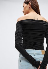 Zwarte off-shoulder top met lange, geplooide mouwen, gemaakt van zacht materiaal. Gezien vanaf de achterkant, gedragen met lichtblauwe spijkerbroek.
