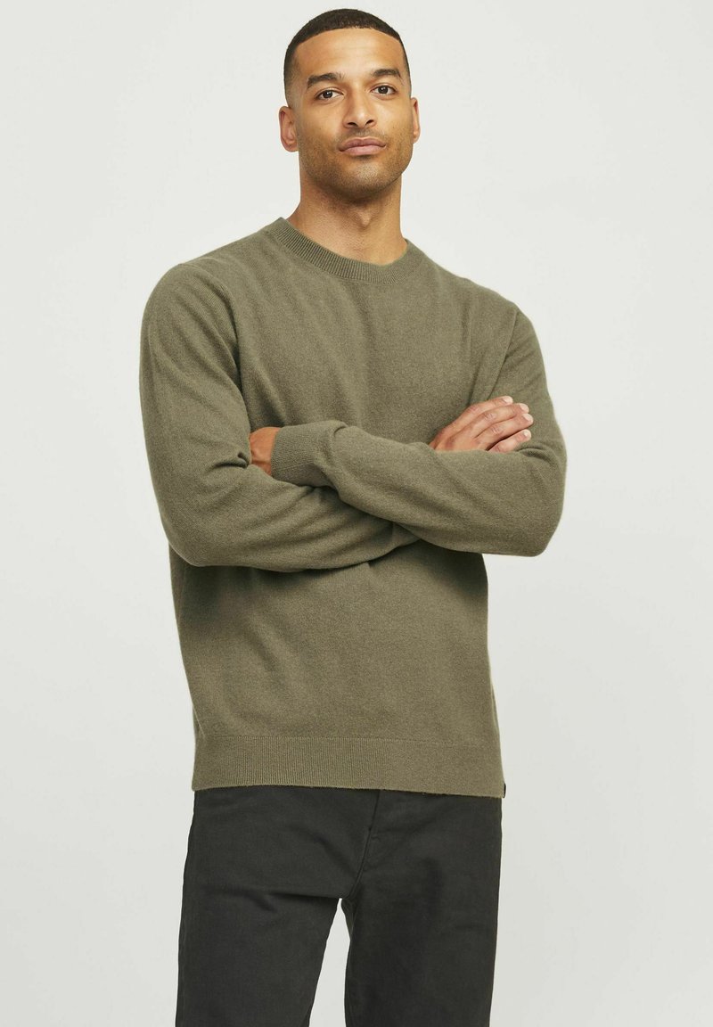 Pulover verde olive dintr-un material tricotat moale, cu decolteu rotund și manșete cu model, asortat cu pantaloni închiși. Textură fină și design simplu.