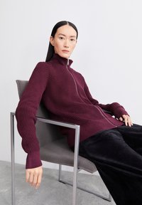 Bordeaux geribbelde zip-up trui met hoge kraag en verlengde mouwen, gecombineerd met donkere broek, zittend op een metalen stoel.