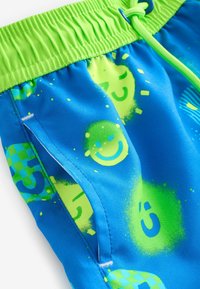 Svøm shorts i klart blåt materiale med grøn elastisk linning. Har blandede grafikker, herunder smilende ansigtter, former og ternede mønstre.