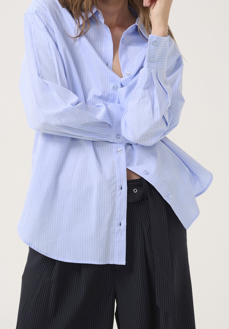 Personne portant une chemise ample à fines rayures bleu clair et un pantalon noir à fines rayures, debout avec un bras plié et la main près du visage.