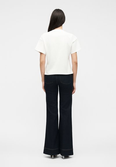 Γυναίκα με μακριά σκουρόχρωμα μαλλιά, που φοράει απλό λευκό κοντομάνικο τοπ και ψηλόμεσα σκούρα μπλε παντελόνια φαρδιάς γραμμής, που στέκεται γυρισμένη με την πλάτη.