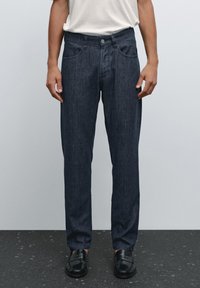 Una persona sta in piedi indossando una maglietta bianca semplice e pantaloni in denim scuro, abbinati a scarpe nere, su uno sfondo neutro.