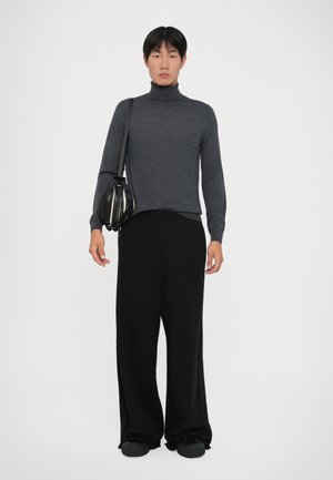 Giovane che indossa un dolcevita grigio scuro, pantaloni neri larghi, scarpe nere e porta una borsa nera a tracolla.