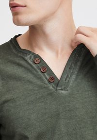 Olajzöld pamut ing V-nyakkal, barna gombdíszítéssel és kontrasztos varrásokkal a nyakrészen. Puha textúra.