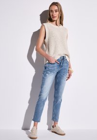 Beige bezrukávový pletený top, světle modré džínové kalhoty s opotřebovaným vzhledem a béžové tenisky s texturovaným designem. Model stojí před jednobarevným pozadím.