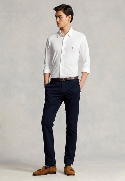 Jeune homme debout les mains dans les poches, portant une chemise blanche boutonnée, un pantalon bleu marine, une ceinture marron et des mocassins marron avec des chaussettes vertes.