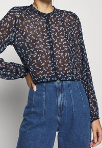 Schwarze durchsichtige Bluse mit kleinem blauem Blumenmuster, Knopfleiste vorne, runder Ausschnitt und langen Ärmeln, kombiniert mit hochgeschnittenen blauen Jeans.