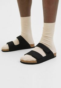 Sorte sandaler med to justerbare stropper, korkfoderseng og struktureret gummisål, iført med lys beige ribbede sokker.