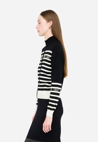 Maglione con zip a righe bianche e nere con colletto alto. Presenta righe strutturate e dettagli con logo sulle maniche. Materiale in maglia liscia.