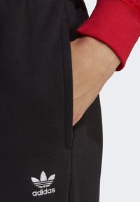Black sports pants with a side pocket, featuring a white Adidas logo. The cuff of a red sweatshirt is visible at the wrist.