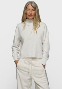 Mujer con cabello largo y gris que lleva un suéter de cuello alto gris claro y pantalones deportivos a juego con cordón, manos en los bolsillos, fondo neutro.