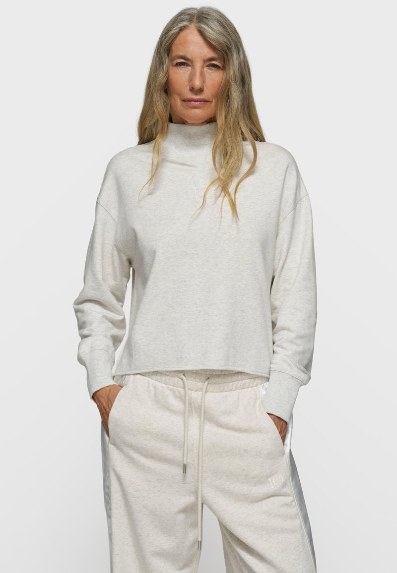 Mujer con cabello largo y gris que lleva un suéter de cuello alto gris claro y pantalones deportivos a juego con cordón, manos en los bolsillos, fondo neutro.