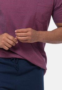 Mains jointes devant une personne portant une chemise maroon rayée et un pantalon bleu marine. L'arrière-plan est blanc uni, mettant l'accent sur les vêtements.