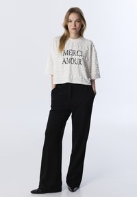 Top corto bianco testurizzato con la scritta "MERCI AMOUR" stampata in nero, abbinato a pantaloni neri a vita alta e scarpe nere eleganti.