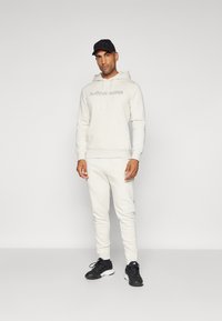 Light beige hoodie with front pocket and "BJÖRN BORG" logo, paired with matching tapered sweatpants and black athletic shoes.