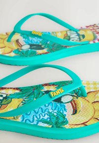 Ryškios turkio spalvos šlepetės su tropiniais pėdų kilimėliais, vaizduojančiais palmes, saulę ir tukano paukštį, pažymėtos geltonu „Fanta“ logotipu.