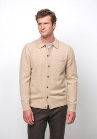 Beige Strickjacke aus Wolle mit Knopfleiste, Kragen und einer Brusttasche. Mit gerippten Manschetten und Saum sowie braunen Knöpfen.