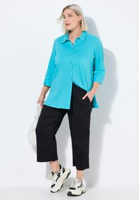 Turquoise knoopshirt van textuurstof, gecombineerd met zwarte cropped broeken. Het model draagt chunky zwart-witte sneakers en houdt een witte tas vast.