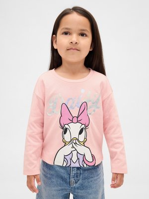 Jauna meitene, tērpusies rozā ar garām piedurknēm kreklā ar lielu Deizi Pīles attēlu un zilās džinsu biksēs, stāv pie vienkārša fona.