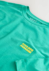 T-shirt turquoise à manches courtes avec texte brodé jaune « POOLSIDE PARADISE » sur la poitrine et étiquette « NEXT » à l'intérieur du col.