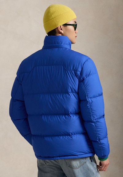 Modra puhasta jakna z visokim ovratnikom in prešito teksturo, ki ima žep z zadrgo, združena z rumeno pleteno kapo in sončnimi očali.