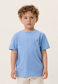 Νεαρός αγόρι με σπαστά ξανθά μαλλιά, ντυμένος με ένα ανοιχτό μπλε κοντομάνικο πουκάμισο και μπεζ βερμούδα, στέκεται μπροστά σε έναν ουδέτερο φόντο.