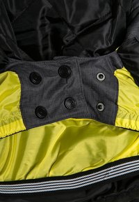Intérieur d'une veste matelassée noire avec une doublure jaune vif. Présente des boutons noirs et un ourlet rayé. Tissu texturé avec des accents lisses.