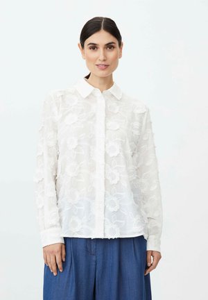 Frau mit dunklem Haar, die eine weiße, langärmelige, mit Blumen bestickte Bluse und blaue, hoch taillierte Hose trägt, steht vor einem schlichten Hintergrund.