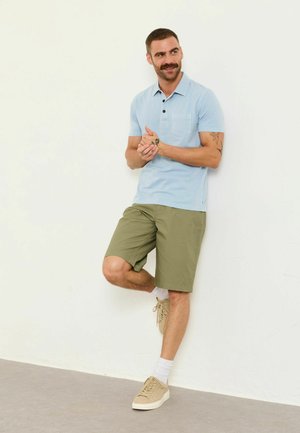 Mann mit Schnurrbart und Tätowierungen, der ein hellblaues Poloshirt, olivfarbene Shorts und beige Sneakers trägt, steht und lehnt an einer weißen Wand.