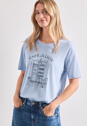 Jeune femme aux cheveux blonds portant un t-shirt bleu clair avec un croquis noir d'une devanture de café et un jean bleu, les mains dans les poches.
