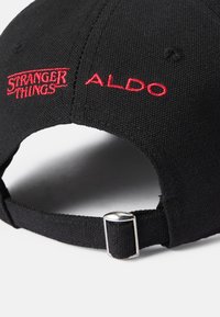 Melnā cepure, kas izgatavota no auduma, ar sarkaniem izšūtiem logotipiem "STRANGER THINGS" un "ALDO." Aizmugurē esošais regulējamais siksniņš ar sudraba metāliskām detaļām.