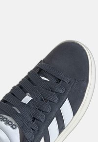 Sivi nubuk tenisice s bijelim bočnim prugama, ravnim sivim vezicama i gumenim potplatom. Imaju teksturirani materijal i šivane detalje.