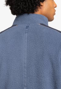 Niebieska bluza polarowa z teksturowanym materiałem, wysokim kołnierzem i zamkiem błyskawicznym. Posiada pionową wstążkę wzdłuż pleców, która dodaje interesującego akcentu projektowego.