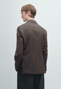 Blazer marrone su misura con colletto a revers, chiusura con un bottone e una texture liscia. Cuciture visibili e un orlo leggermente curvo.