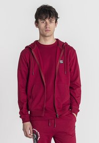 Červená zipsová mikina s predným vreckom, vyrobená z mäkkého materiálu. Kombinovaná s farebne zladeným červeným tričkom a nohavicami s grafickým dizajnom.