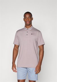 Hnedý polo tričko s krátkym rukávom a golierom, so zapínaním na tri gombíky a čiernym logom Nike na ľavej hrudi, nosené s modrými vzorovanými šortkami.