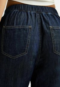 Pantalons en denim foncé avec un waistband élastique, dotés de deux poches arrière et de coutures contrastées. Le tissu a une texture douce.