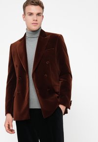 Jaunas vyras, vilkintis rudą aksominį dvigubos sagos švarką ant pilkos golfinės megztinio ir juodų kelnių, stovi prieš baltą foną.