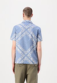 Camicetta azzurra a maniche corte con motivi geometrici e paisley bianchi, dotata di colletto a punta e vestibilità morbida; realizzata in tessuto leggero.