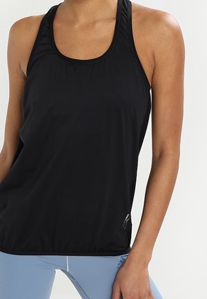 Kobieta ubrana w czarny, sportowy, bezrękawnik i jasnoniebieskie legginsy na jednolitym tle.