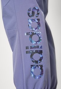 Pantalon de sport violet en tissu doux, arborant un grand logo "adidas" coloré au design pixélisé sur le côté.