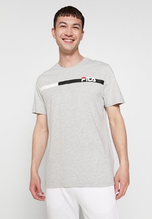 Homme souriant portant un t-shirt FILA gris clair avec des rayures noires et blanches sur la poitrine, associé à un pantalon blanc, debout devant un fond blanc.