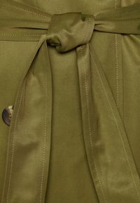 Olijfgroen stof met een gladde textuur heeft een gevouwen strik rond de taille, vastgemaakt in een knoop, en is geaccentueerd met zichtbare stiksels.