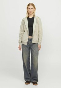 Haut à capuche zippé gris clair avec une capuche, des poignets côtelés et des poches kangourou, associé à un jean en denim sombre ample et des bottes marron.