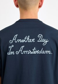 Navyn blå bomull T-skjorte med hvit, kursiv tekst på baksiden som sier "En dag til i Amsterdam..."