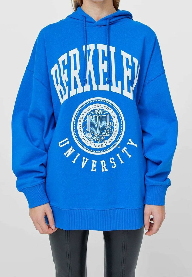 Personne portant un sweat à capuche bleu vif de l’Université de Berkeley avec texte et emblème blancs, associé à un pantalon noir sur un fond clair.