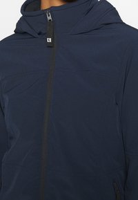 Námořnicky modrá bunda s kapucí vyrobená z lehkého materiálu, s předním zipem, bočními kapsami a minimalistickým designem s jemnými švy.