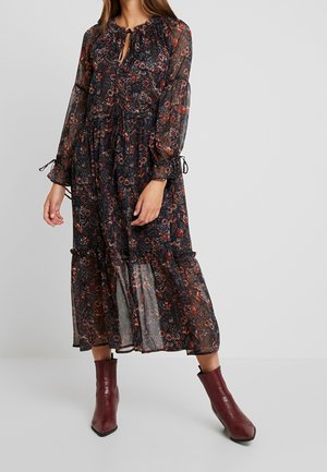 Frau trägt ein langärmeliges schwarzes Kleid mit Blumenmuster aus transparentem Stoff und bordeauxrote Stiefeletten vor schlichtem grauem Hintergrund.