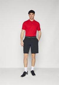 Crvena polo majica, crne hlače s remenom, bijele čarape i crne tenisice. Model stoji protiv jednostavne pozadine.